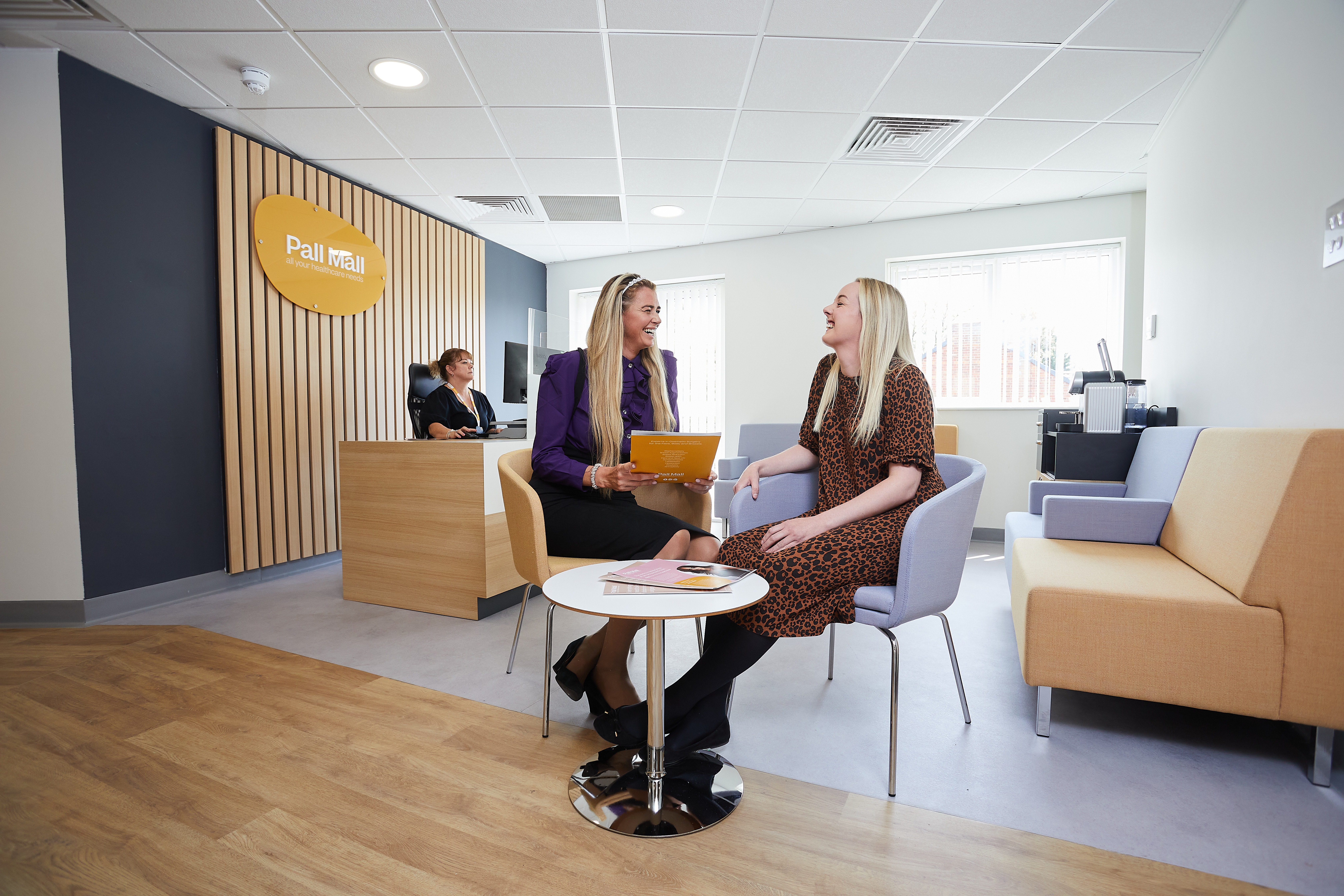 Patient coordinator with patient sat at a table in waiting area laughing Patient coordinator with patient sat at a table in waiting area laughing
